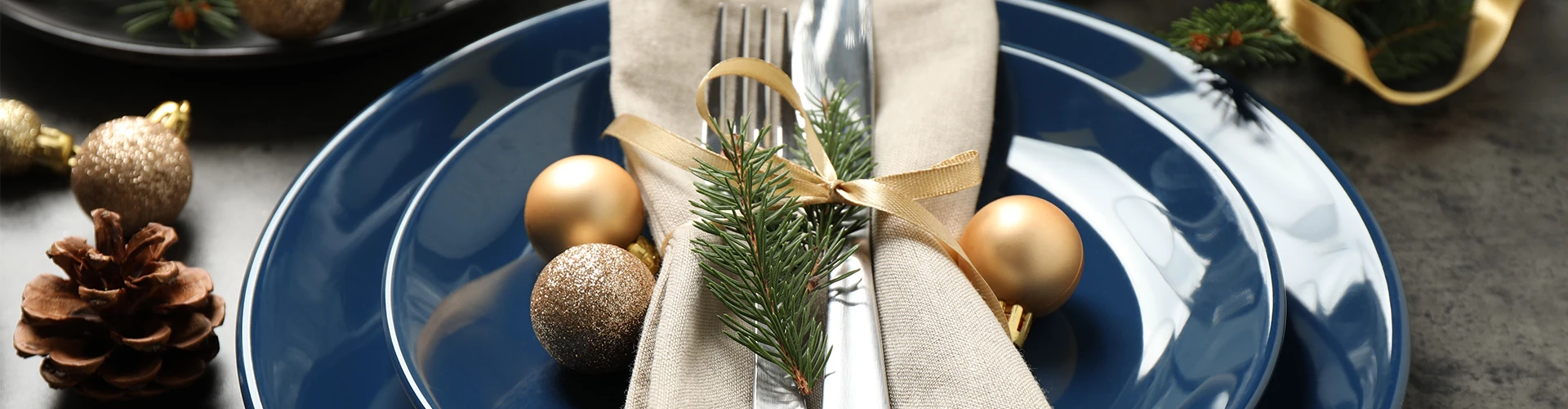 Een feestelijk gedekte tafel met een blauw bord, gouden kerstballen, dennenappel, en bestek gewikkeld in een beige servet met dennentak.
