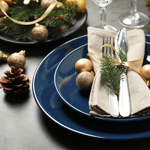 Een feestelijk gedekte tafel met een blauw bord, gouden kerstballen, dennenappel, en bestek gewikkeld in een beige servet met dennentak.