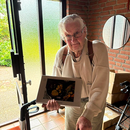Dhr. Meershoek met bril en rollator houdt een puzzeldoos vast bij een deur met matglas, tegen een bakstenen muur.