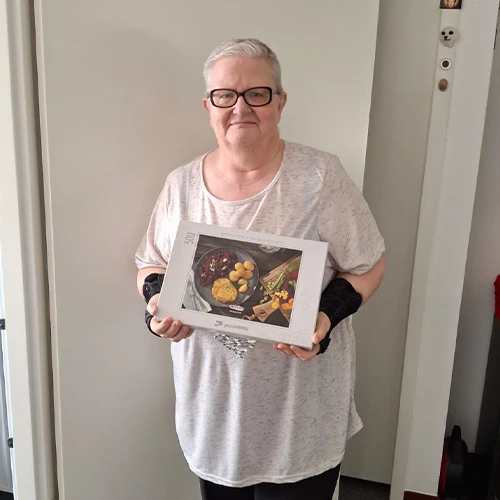 Mevr. Heijnen in lichtgekleurd shirt en zwarte bril houdt een puzzeldoos vast in haar woonkamer.