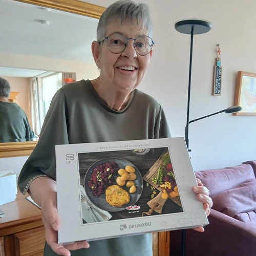 Glimlachende mevr. Raaymakers met bril die een puzzeldoos vasthoudt in een woonkamer.
