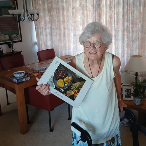 Lachende mevr. Segers met wit haar en bril, bij rollator, houdt een 500-delig puzzeldoosje vast in een woonkamer.