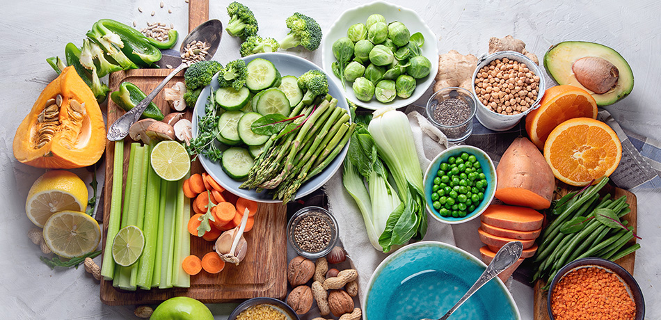 Verschillende verse groenten en fruit, waaronder pompoen, komkommer, spruitjes, sinaasappel, avocado en peultjes, op een tafel.
