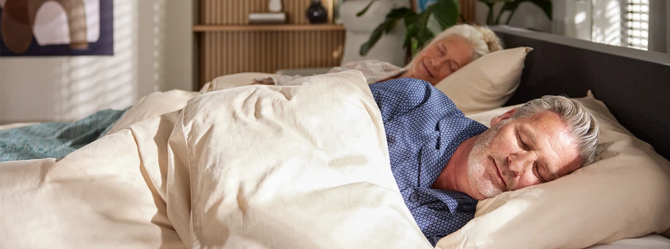 Een ouder stel slaapt vredig in een goed verlichte slaapkamer, beiden met een ontspannen uitdrukking op hun gezicht. Het bed heeft een antraciet hoofdbord en beigekleurige lakens.