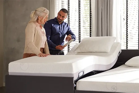 Een oudere vrouw en een man bespreken een verstelbaar bed in een showroom. Het bed heeft twee afzonderlijk verstelbare matrassen.