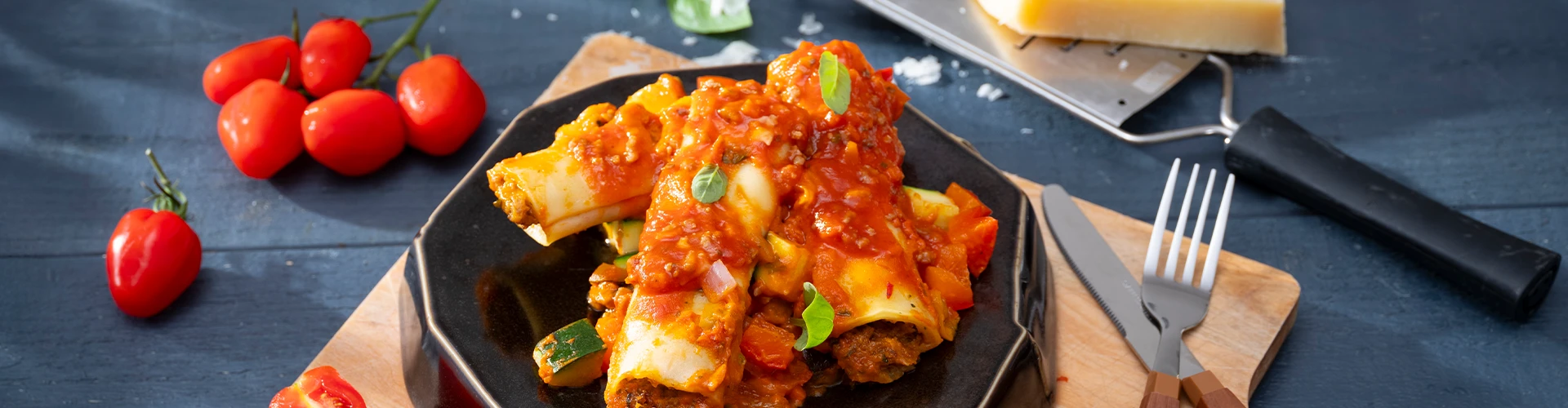Cannelloni met gehakt, groenten en tomatensaus, gegarneerd met basilicum, op een zwart bord, geplaatst op een snijplank met een vork, mes, kaasrasp en een blok Parmezaanse kaas ernaast.