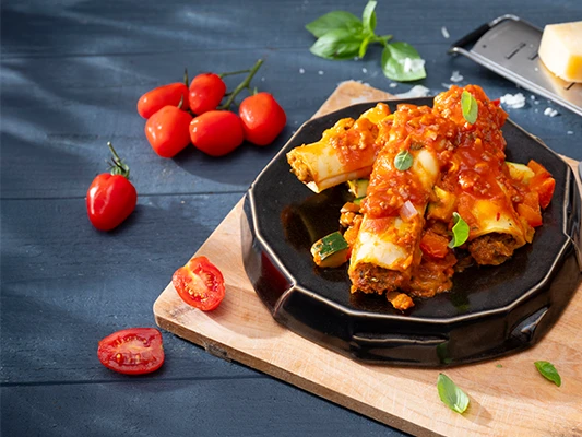 Cannelloni met gehakt, groenten en tomatensaus, gegarneerd met basilicum, op een zwart bord, geplaatst op een snijplank met een vork, mes, kaasrasp en een blok Parmezaanse kaas ernaast.