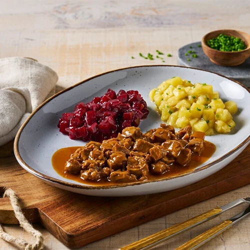 Een lichtblauw bord met gestoofd rundvlees in jus, met rode bieten en aardappelen in saus. Gesneden in kleine blokjes van 1,5 cm. Geserveerd op een houten plank met een potje verse kruiden.