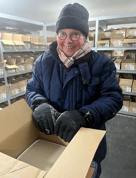 Vrouw met bril en warme jas, muts en handschoenen glimlacht terwijl die dozen inpakt in een koude opslagruimte.