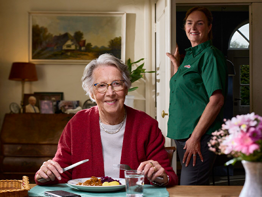 Bezorger van apetito kijkt bij senior vrouw aan de eettafel met haar maaltijd