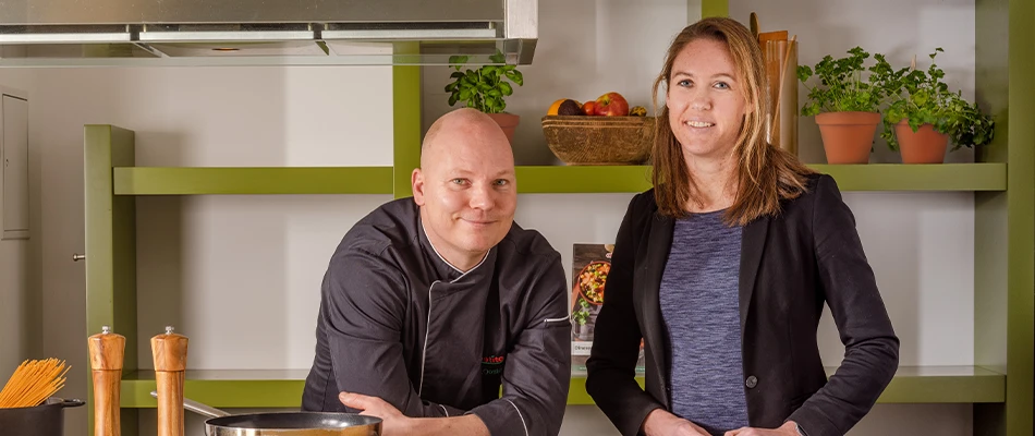 Chefkok Chris in zwarte koksbuis leunend aan keukenwerkblad naast diëtist Karin in zwarte blazer. Kruiden en keukengerei op planken achter hen.